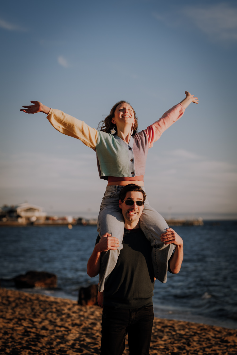 Couple at their Engagement Session by the beach. She is riding on his shoulders with her hands in the air while he pulls a funny face.