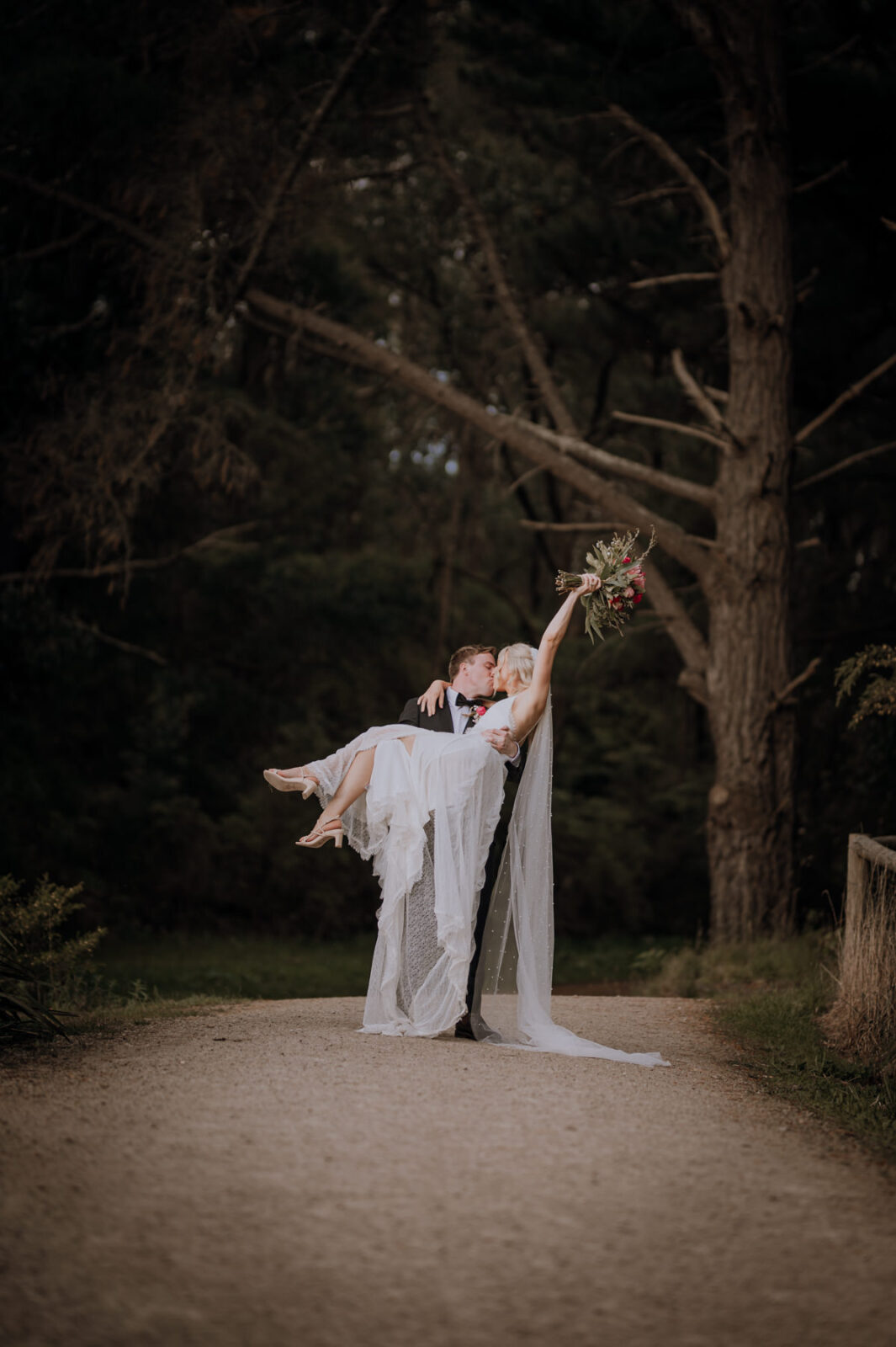 Groom carrying bride on a path in the forest, bride is kissing groom with bouquet raised in the air.