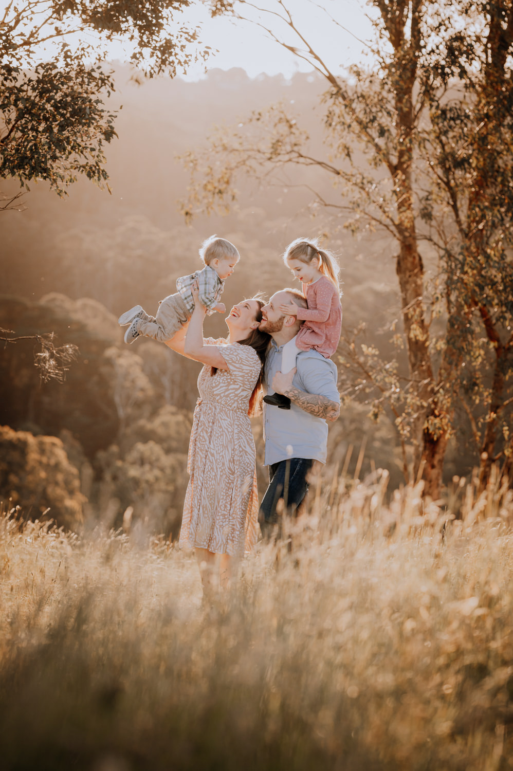 Family of four standing amidst golden grass, mum is holding little boy up in the air and the little girl is sitting on Dad's shoulders.