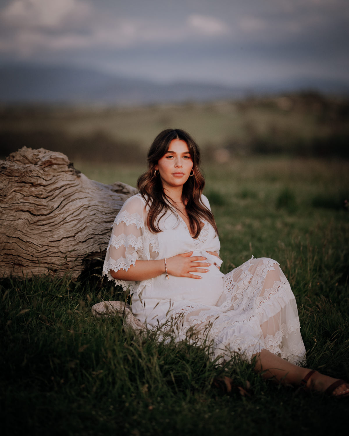 Pregnant lady at her maternity session sitting in the grass leaning against a fallen log with hills in the distance behind her.