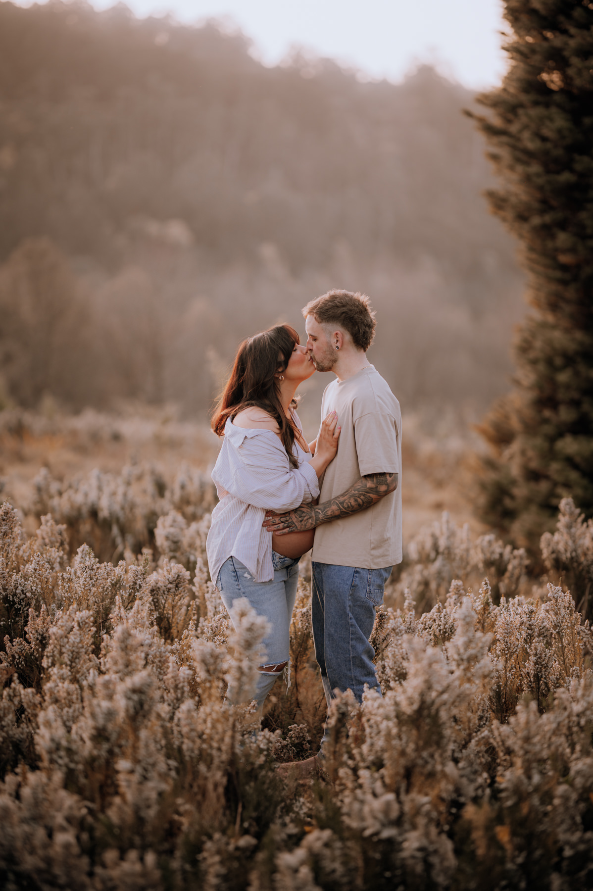 Photo from a Mt Dandenong Maternity session. A couple are kissing in the middle of a field of heather in the mountains. She is pregnant and the scene is awash with golden light.